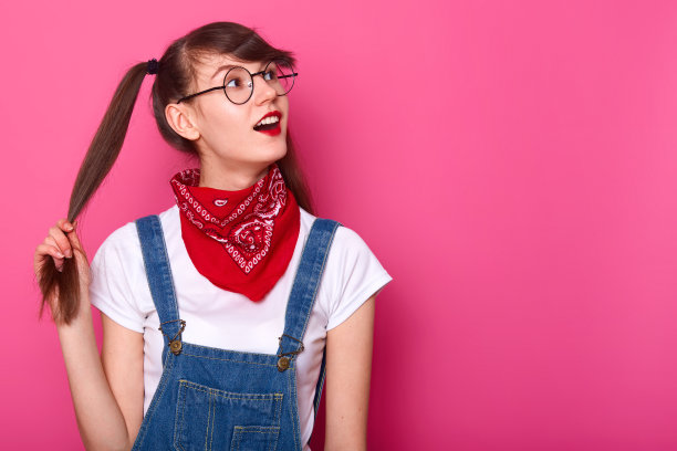 年轻的女性，有着健康闪亮的棕色头发，手里拿着马尾辫。女同学对什么东西感兴趣，张着嘴看着左上角。模特在工作室里靠着粉红色的墙摆姿势。图片下载