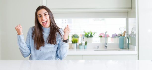 广角图片美丽的年轻女子坐在白色的桌子在家里庆祝惊讶和惊讶的成功与双臂举起和睁开眼睛。赢家的概念。图片下载