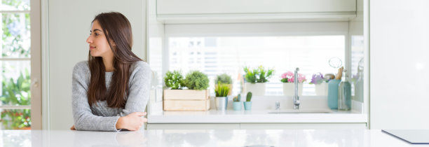 广角图片美丽的年轻女子坐在白色的桌子在家里看向一边，放松的侧面姿势与自然的脸与自信的微笑。图片下载