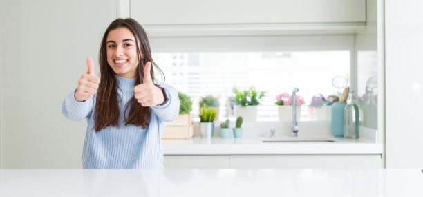 广角图片美丽的年轻女子坐在白色的桌子在家里成功的手势与手做积极的手势，拇指向上微笑和幸福。带着愉悦的表情看着镜头，胜利者的姿态。图片下载