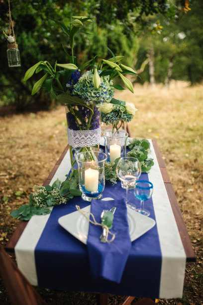装饰。细节。婚礼装饰。在树林里的木桌上摆放着一幅鲜花，上面有蓝色、白色、绿色的鲜花和绿叶植物，蜡烛，小雕像，玻璃器皿，餐具。特写照片图片下载