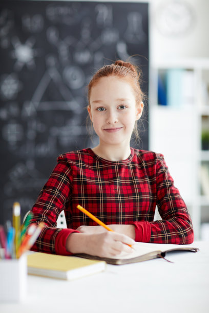 内容漂亮的学生女孩在红色格子裙坐在书桌在学校，看着相机，同时做笔记在工作簿图片下载