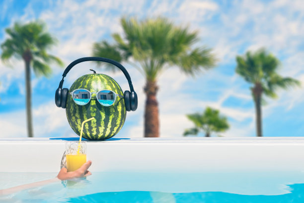 这是一张很有创意的夏日照片，西瓜戴着太阳镜和耳机，在棕榈树上的泳池边用吸管喝果汁。概念图片下载