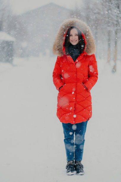这张照片拍摄的是一个穿着红色冬季夹克、牛仔裤和靴子的漂亮女人，双手插在口袋里，在冬天雪花漫天的时候站在街上，摆姿势拍摄户外照片图片下载