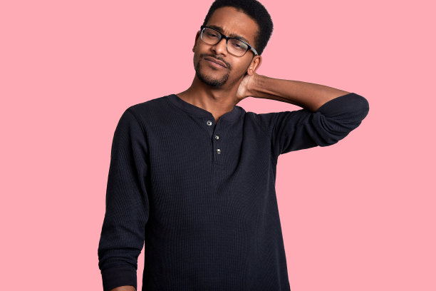 英俊的黝黑皮肤的男性在辛苦工作一天后看起来很累。男人总是把手放在脖子上。年轻人戴着眼镜，穿着黑毛衣。模特在照片工作室的孤立在粉红色的背景。图片下载