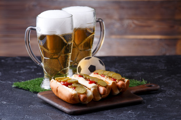 两杯泡沫啤酒的照片，绿茵茵的草地上有足球，热狗图片下载