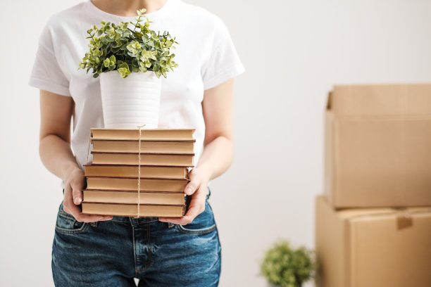 搬迁的概念和搬到一个新的家。特写中，女性的双手捧着一堆书和一盆绿色植物。背景是纸板箱。图片下载