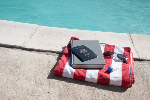 美国护照，小说，太阳镜，旅行装的防晒霜放在红白条纹的沙滩浴巾上，旁边是游泳池。游泳池静物，准备去度假。图片下载