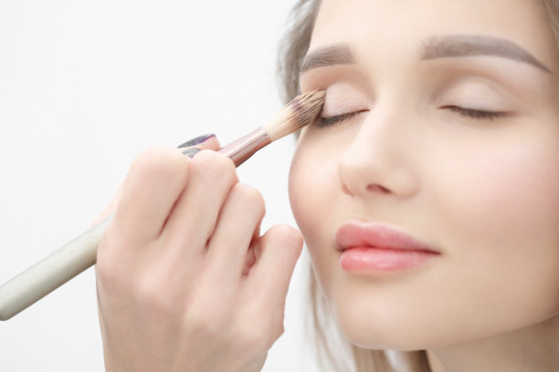 化妆师在白色的背景上化妆美丽的金发女孩。特写手与刷子和脸。图片下载