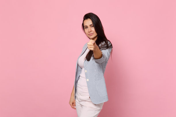 微笑的年轻女子的肖像在条纹夹克指着食指在相机孤立的粉色粉彩墙背景在工作室。人真诚情感，生活理念。模拟拷贝空间。图片下载