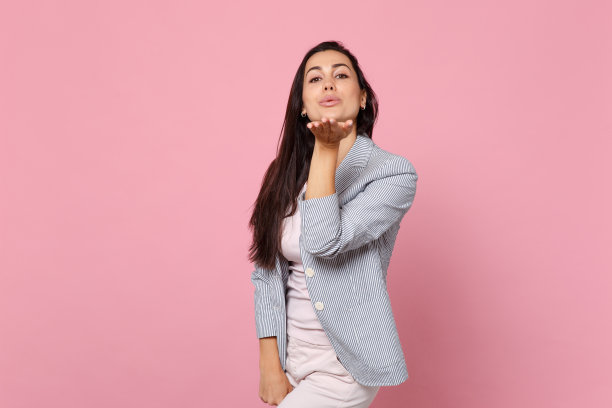 肖像温柔美丽的年轻女子在条纹夹克吹，送空气亲吻孤立在粉红色的粉彩墙背景在工作室。人真诚情感，生活理念。模拟拷贝空间。图片下载