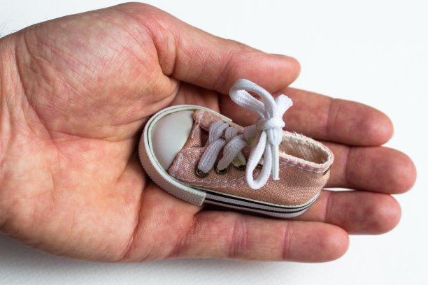 男人的大而有力的手里拿着一双小巧的童鞋。鞋上系着一个蝴蝶结。一只手拿着鞋子放在白色的背景上，突出了它们。图片下载