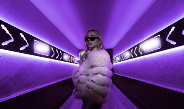 青少年潮女孩在时尚的眼镜和皮草站在紫色霓虹灯元素在大街上的墙，女青少年时尚模特在城市夜晚紫色明亮的俱乐部发光，回到80年代图片下载