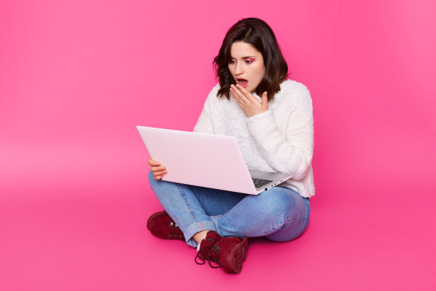 美女睁大眼睛看着笔记本电脑屏幕，双腿交叉坐在地板上，手放在嘴上。女孩在社交网络上看到了令人震惊的闺蜜和男友的照片。图片下载