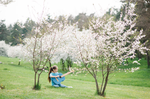 美丽的小女孩弹着原声吉他，唱着歌，坐在春天公园里盛开的树木下的绿草地上图片下载