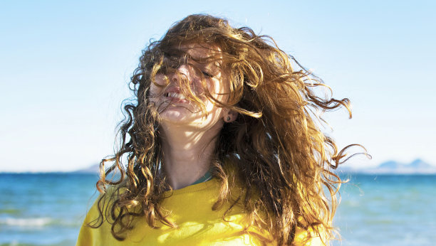 一个有风的日子，美丽的年轻女子在海滩上放松。快乐的金发女孩对着海景微笑。图片下载