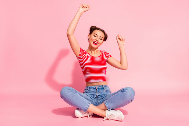 全身的身体尺寸照片美丽的她，她的女士发髻，润发油，嘴唇，双手，双臂，空中，眼睛，闭上，拉拉队队长，穿着休闲牛仔裤，牛仔，条纹，红色，白色t恤，坐着，地板，孤立的，粉红色的背景图片下载