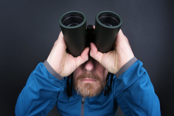 在灰色的背景下，蓄着胡子的人用双筒望远镜抬头看图片下载