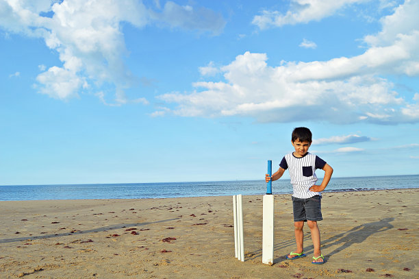 水平的照片，一个小男孩站在沙滩上，拿着一个板球拍和三柱门在他的身边在一个夏天。图片下载
