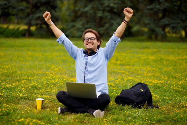 一名年轻男子在公园里心情愉快，成功地举起双手，在外面用手提电脑。教育的概念。图片下载