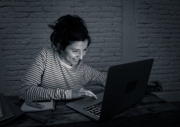 近距离拍摄的黑白肖像，一个快乐随意美丽的女人工作和学习在笔记本电脑上坐在她的桌子晚上。在家工作和上网的概念。图片下载