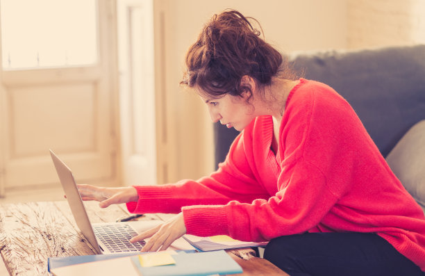 一个深思的随意美丽的女人穿着明亮的红色套头衫工作和学习在电脑上坐在家里的沙发上。在家工作和上网的概念。图片下载