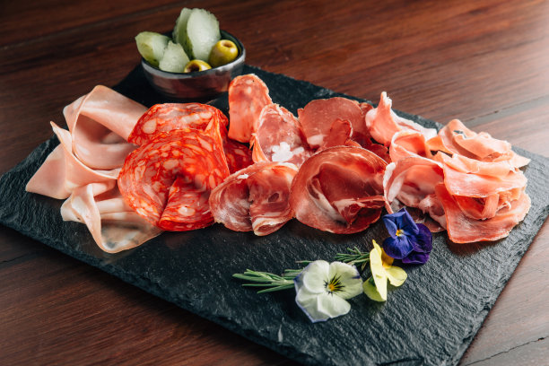 石盘上的冷切肉，配以意大利熏火腿、熏肉、意大利腊肠和香肠，并以花朵装饰在木质背景上。肉拼盘开胃菜与泡菜和橄榄。图片下载