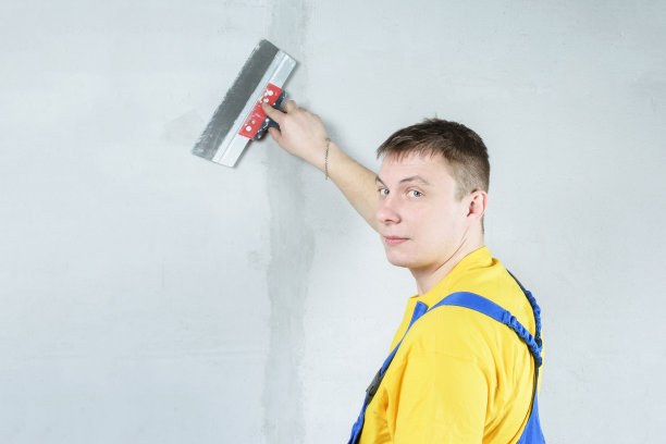 建筑工用泥铲小心地把墙上不平整的地方修整好。穿着工作服的建筑工靠着一堵灰色的墙。照片抹灰工作，与文本空间。图片下载