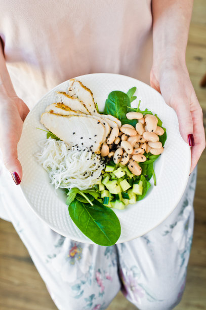 女孩端着一个佛碗，里面有面条、豆子、鸡胸肉、菠菜、芝麻菜和黄瓜。白色背景，俯视图图片下载
