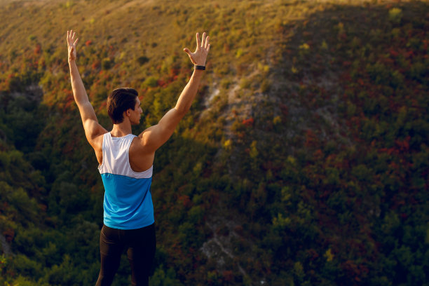 一个运动员的图片，肌肉发达，健康，腹肌，运动的年轻人做伸展运动举起双手，在户外的森林，高山高的背景。夏天的日落景观。图片下载