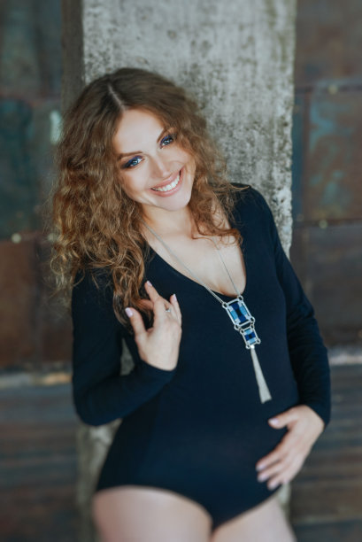 迷人的微笑孕妇半裸女性内衣黑色紧身衣与卷曲的头发和亮蓝色吊坠项链摆姿势在工作室。程式化的深色照片拍摄图片下载