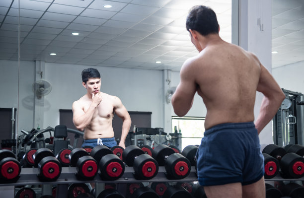 亚洲英俊年轻肌肉发达的亚洲男子在健身房的一个镜子前锻炼健美。一个人弯曲训练他的二头肌，在健身房的镜头前展示身体。图片下载