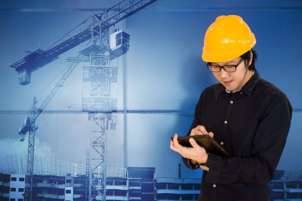 蓝色背景上，亚洲男性工程师戴着防护安全帽，手持数码板，现场施工起重机。土木工程和建筑施工概念图片下载
