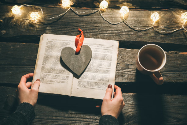 节日气氛的照片。圣诞彩灯和热茶杯。女孩手里拿着一本书，准备度过一个温馨的夜晚。托盘上的木心。完美的冬季平躺。Hygge概念。图片下载