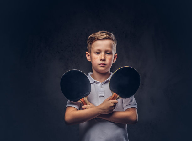 可爱的红发男孩穿着一件白色t恤，双手交叉拿着两个乒乓球拍。孤立在深色纹理背景上。图片下载