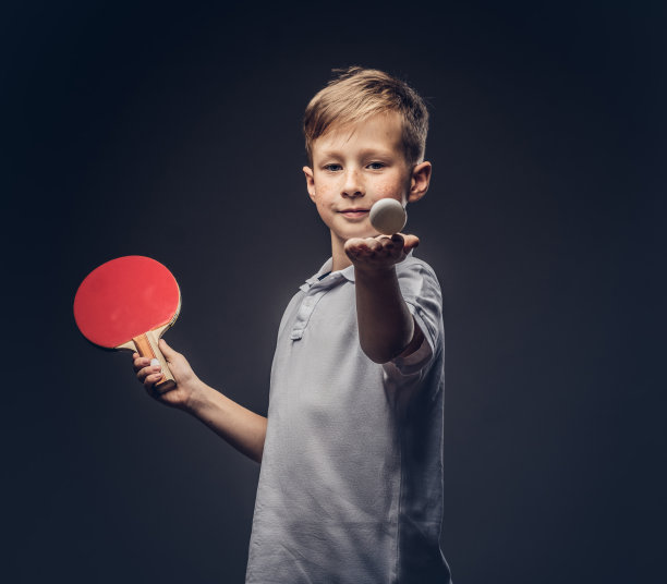 一个红头发的男生穿着一件白色的t恤，拿着一个乒乓球拍和一个球。孤立在灰色背景上。图片下载