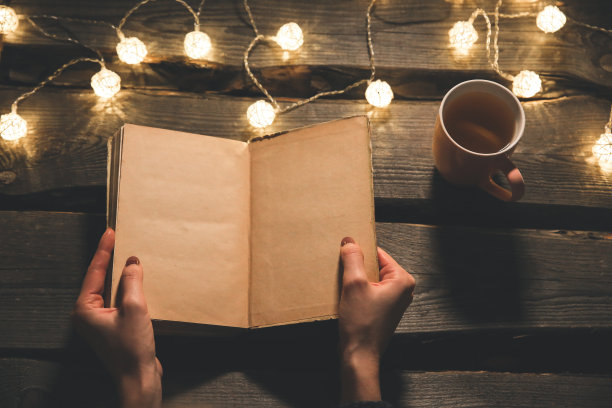 节日气氛的照片。圣诞彩灯和热茶杯。女孩手里拿着一本书，准备度过一个温馨的夜晚。女性阅读。完美的冬季平躺。Hygge概念。图片下载