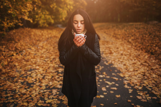 一个年轻的女孩拿着一杯热饮，微笑着看着秋天的森林。迷人的年轻女子在秋天户外。女模特在秋天的森林里喝着热茶，特写图片下载