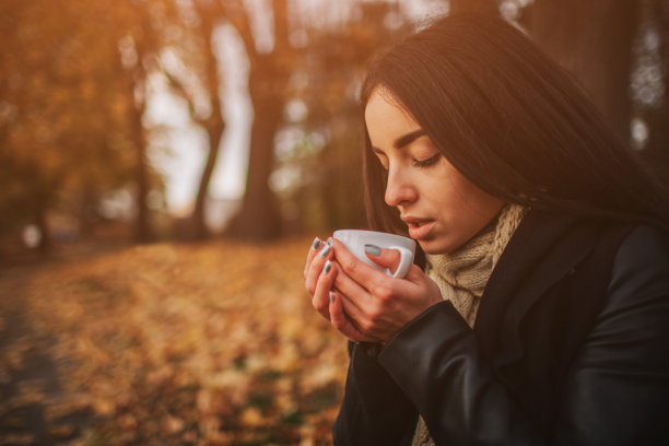 一个年轻的女孩拿着一杯热饮，微笑着看着秋天的森林。迷人的年轻女子在秋天户外。女模特在秋天的森林里喝着热茶，特写图片下载