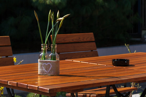 木桌上放着一个带白色心形图案的小玻璃瓶，花朵和花蕾在里面盛开，旁边放着一个黑色的烟灰缸和几把椅子。餐厅露台的简单装饰概念。图片下载