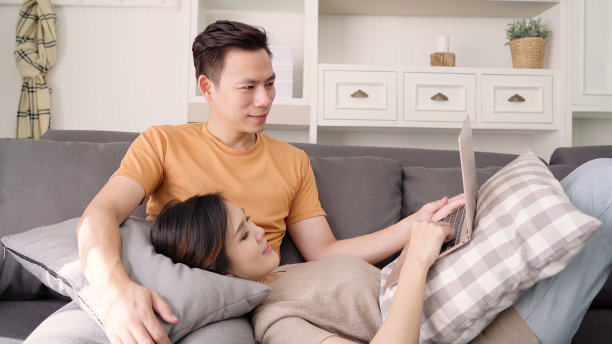 亚洲情侣在家里的客厅里用笔记本电脑搜索网络，甜蜜的情侣在家里放松的时候躺在沙发上享受爱情的时刻。生活方式夫妇在家里放松的概念。图片下载