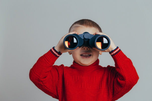 灰色背景中，一个穿着红毛衣微笑迷人的男孩用黑色望远镜看近景图片下载