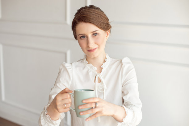 有吸引力的年轻女子在休闲衣服拿着一个杯子，看着别处和微笑，在白色的背景。图片下载