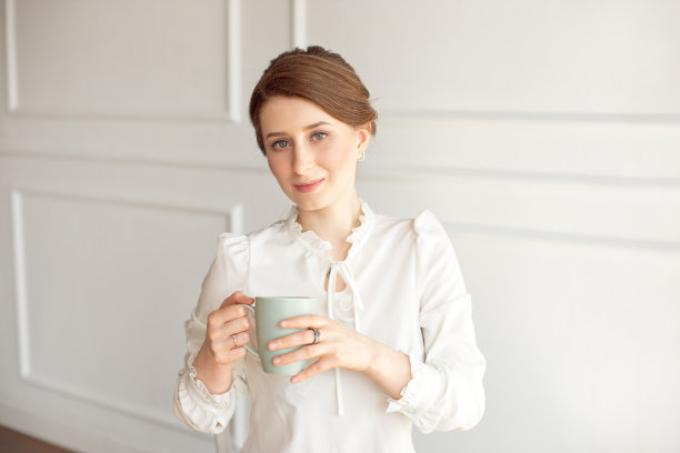 有吸引力的年轻女子在休闲衣服拿着一个杯子，看着别处和微笑，在白色的背景。图片下载