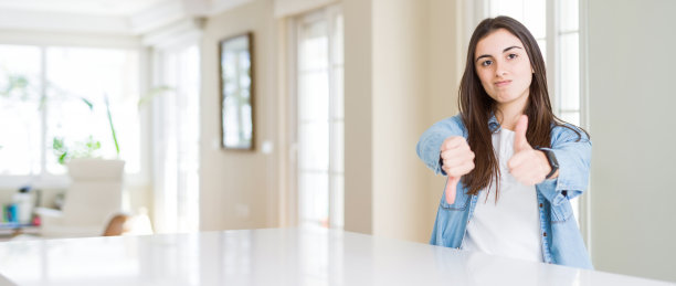 广角图片美丽的年轻女子坐在家里的白色桌子上做拇指向上和向下，不同意和同意的表达。疯狂的冲突图片下载