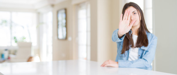 广角图片美丽的年轻女子坐在白色的桌子在家里做停止与手掌唱歌。警告表情与否定和严肃的手势在脸上。图片下载