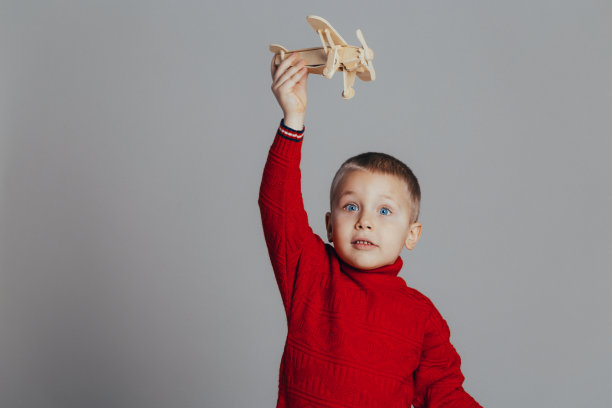一个穿着红色毛衣的帅气男孩在灰色背景上玩木制飞机的特写镜头图片下载