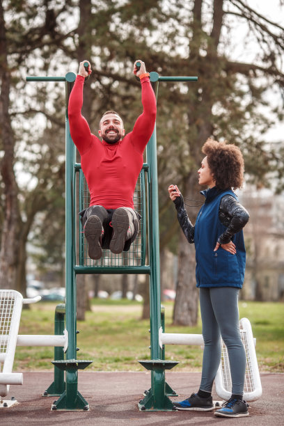 肌肉男在公园的引体向上做下腹肌锻炼，他的私人健身教练站在他旁边，并测量他的表现图片下载