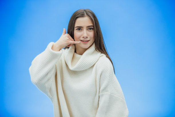 蓝色背景上的漂亮女孩。女孩做了个手势，表示正在打电话。穿着一件白色暖和的毛衣。有黑色的长发。图片下载