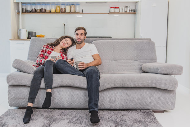 一对漂亮的年轻人。爱的人们捧着杯子，妻子把头靠在她的肩膀上。在看电视的时候。坐在家里的沙发上。图片下载